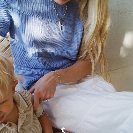 Woman in a blue sweater interacting with a child on a white surface, wearing a cross  pearl necklace and cross bracelet set.