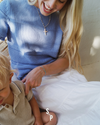Woman in a blue sweater interacting with a child on a white surface, wearing a cross  pearl necklace and cross bracelet set.