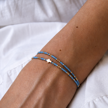 A close-up of a person's wrist wearing two delicate beaded bracelets, one light blue and one gold with a small gold cross, resting on white fabric.