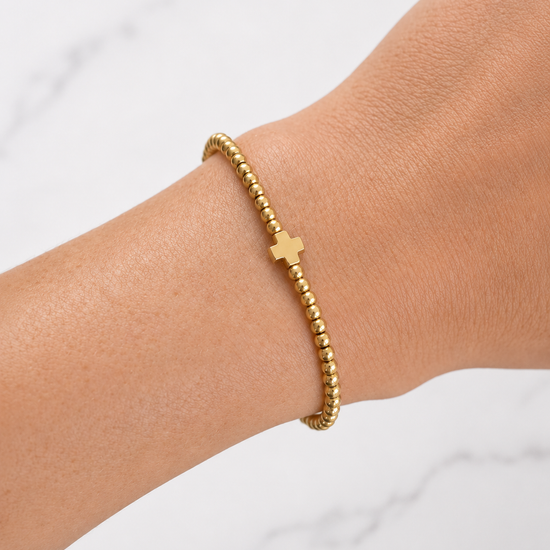 A delicate gold beaded bracelet with a small cross charm, shown worn on a person's wrist against a light marble background.