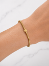 A delicate gold beaded bracelet with a small cross charm, shown worn on a person's wrist against a light marble background.