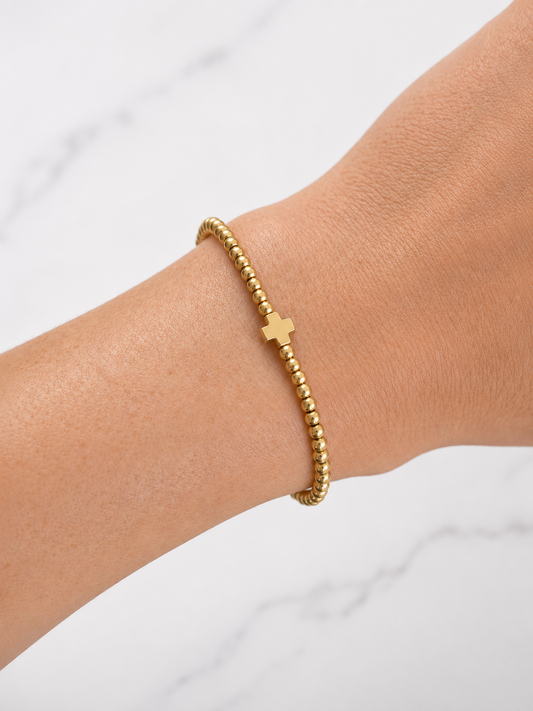 A delicate gold beaded bracelet with a small cross charm, shown worn on a person's wrist against a light marble background.