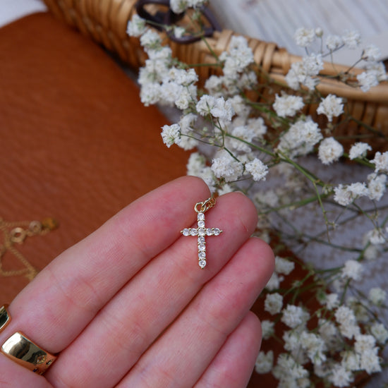 Gold cross pendant held by a hand with small white flowers in the background