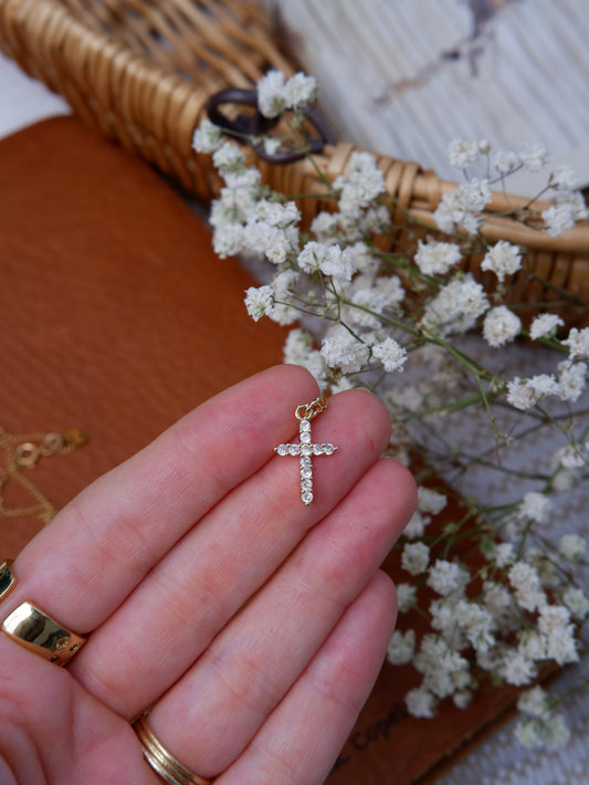 Gold cross pendant held by a hand with small white flowers in the background