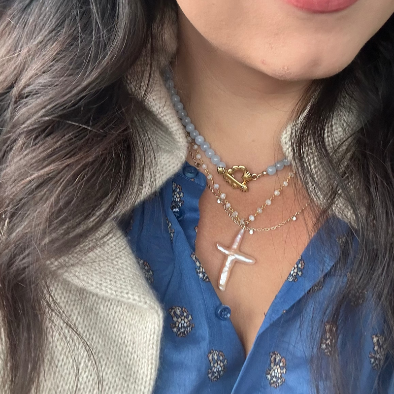 Close-up of a person wearing layered necklaces, including a silver cross pendant, a gold clasp necklace, and a light blue beaded necklace, styled with a blue patterned blouse and a beige knit cardigan.